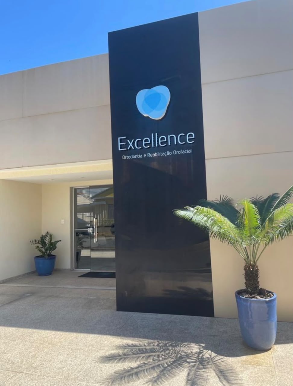 Fachada da Excellence, com letreiro azul, entrada de vidro e vasos junto à porta